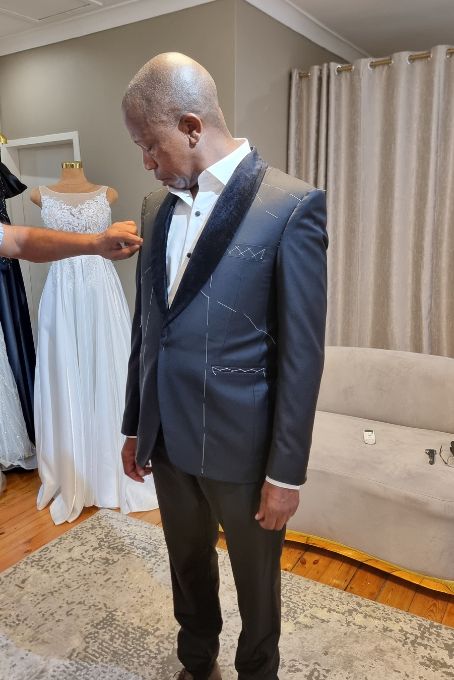 A groom in a navy dinner suit at his fitting, beside a wedding gown